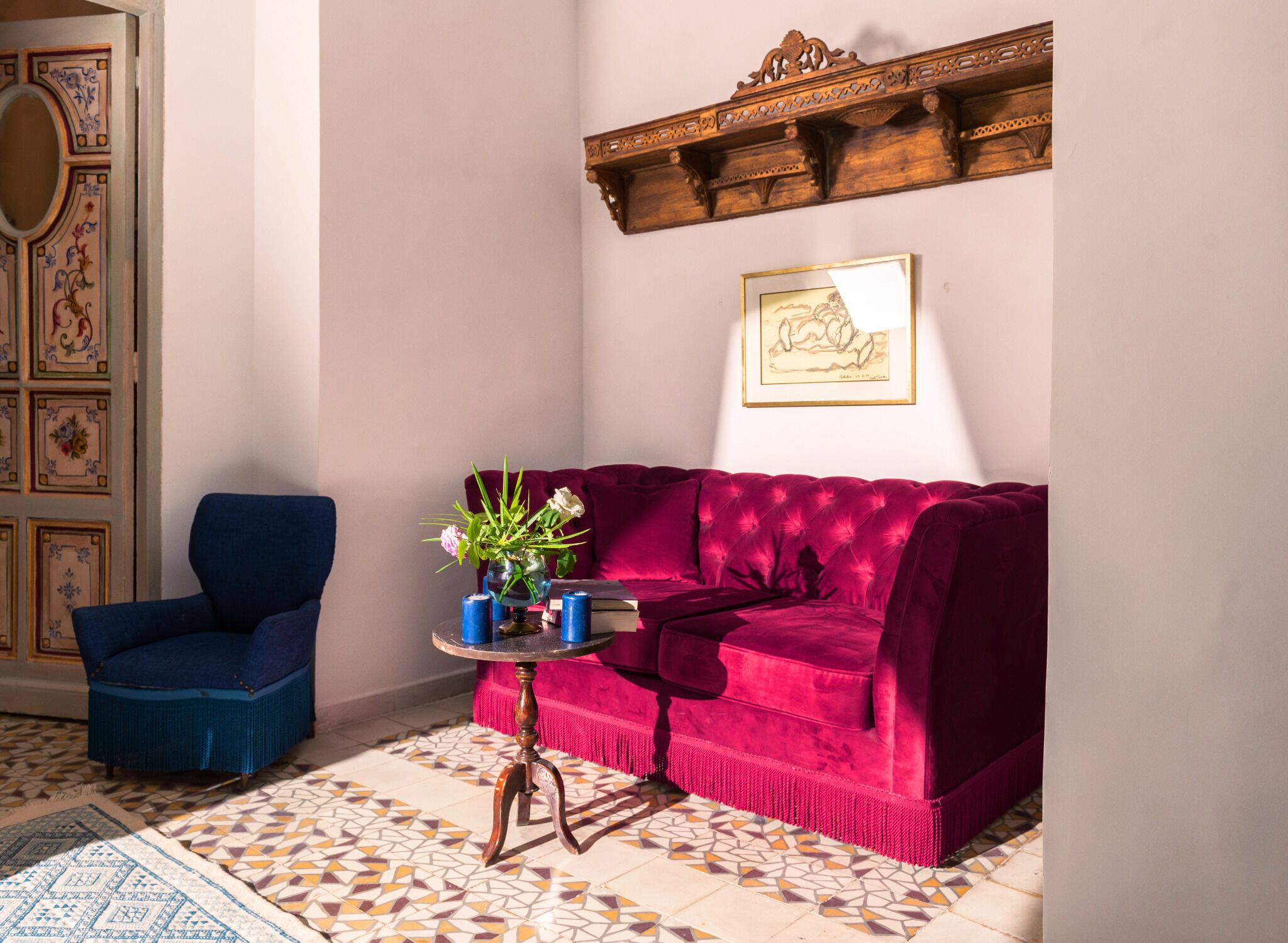 Una stanza con una poltrona blu, un divano rosso, un tavolino con un vaso di fiori e un pavimento decorato.