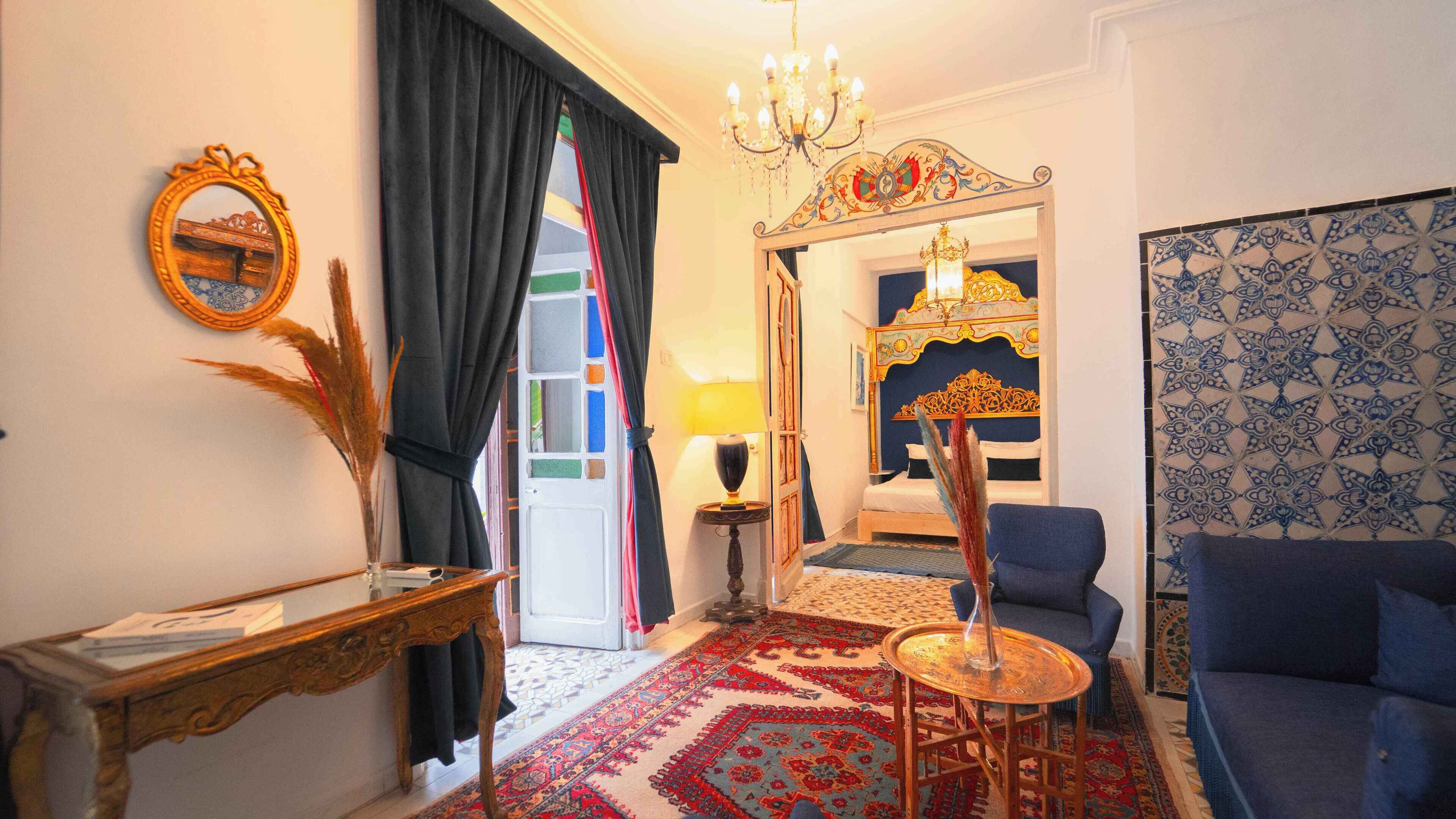 Ein Zimmer im traditionellen Stil mit einem Bett mit aufwendiger Kopfteil, einem blauen Sofa, einem kleinen Tisch mit einer Lampe und einem dekorativen Spiegel.