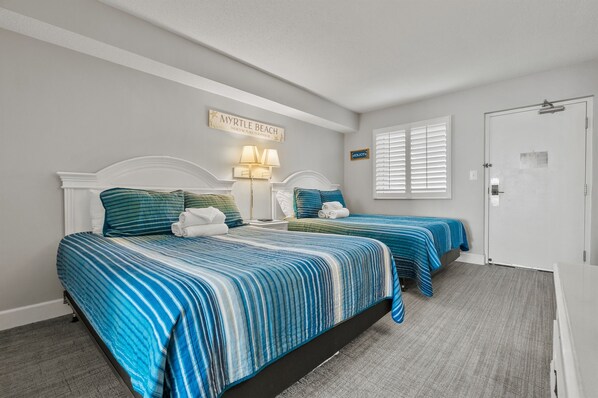 A hotel room with two beds, each with blue-striped bedding, a white headboard, and a wall-mounted lamp.