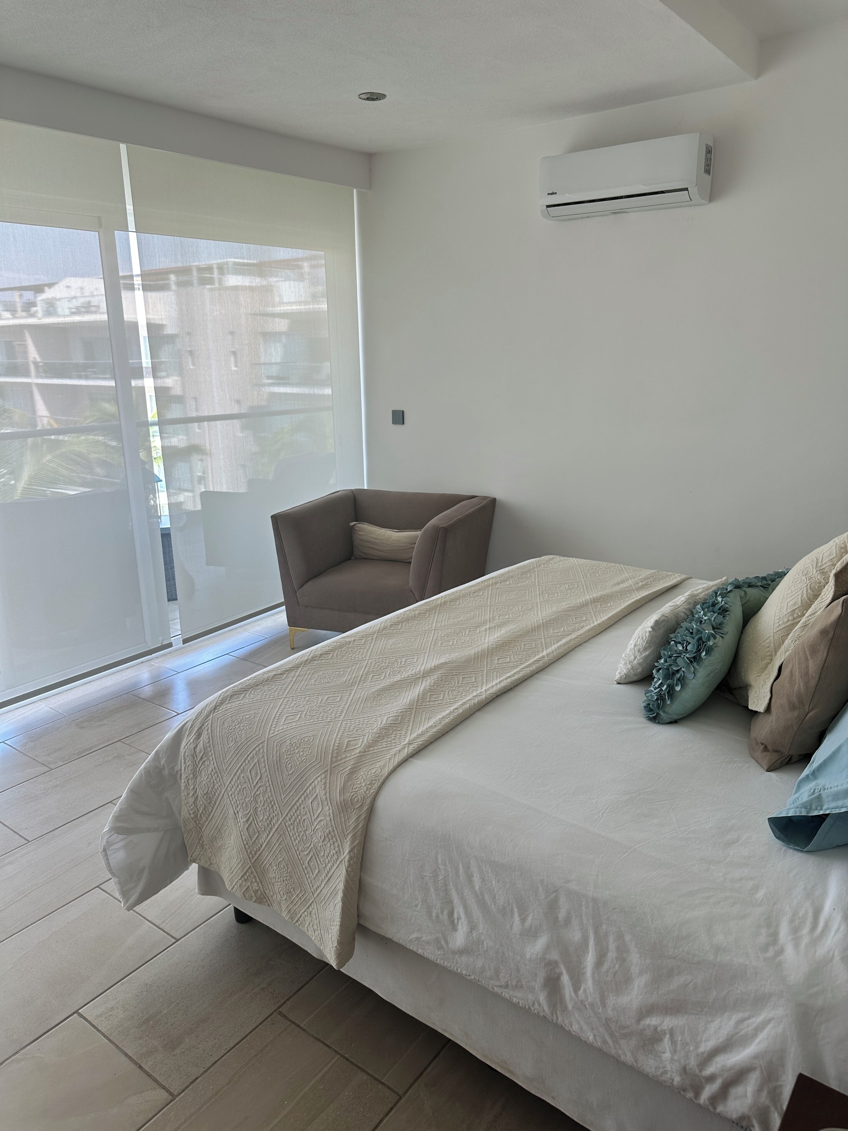 A bedroom with a large bed, a chair, a window, and an air conditioning unit.