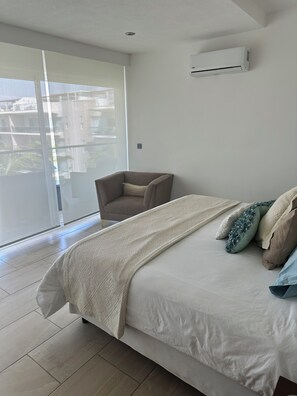 A bedroom with a large bed, a chair, a window, and an air conditioning unit.