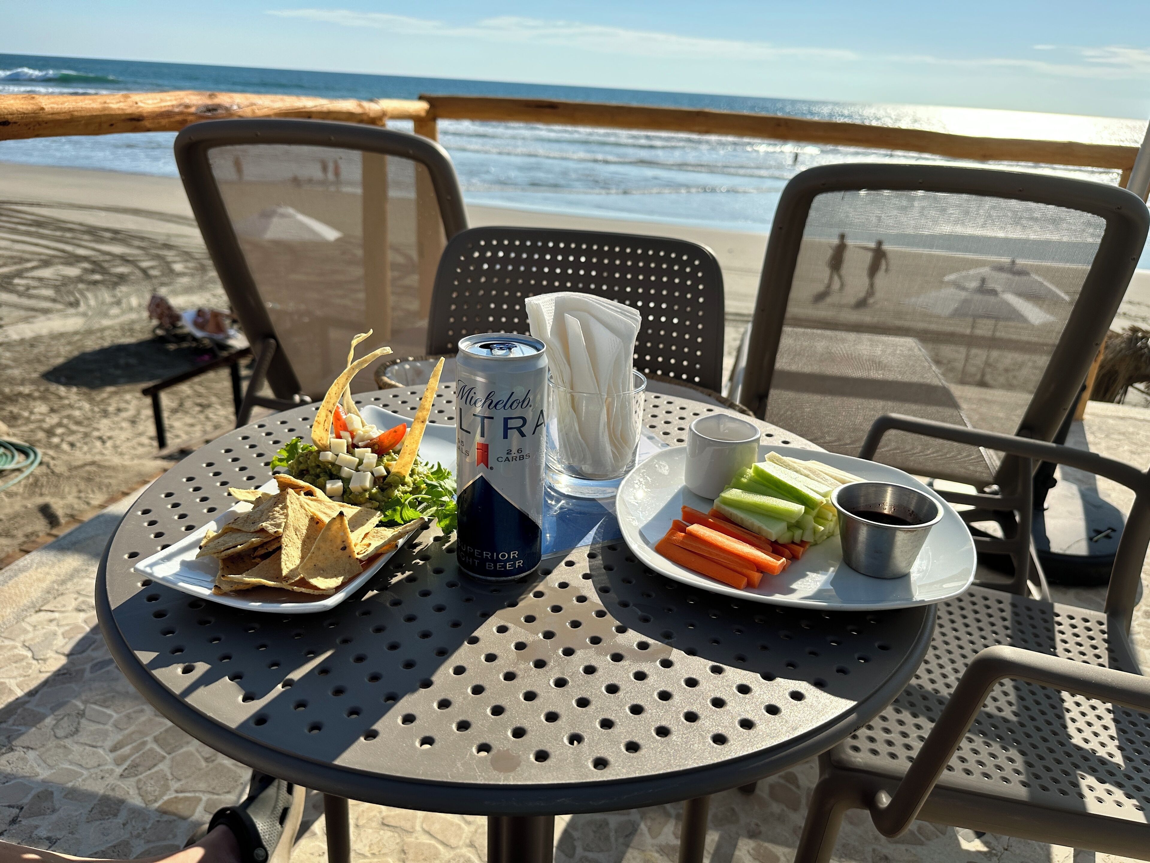 A table with a can of beer, a plate of vegetables, and a glass of water on a beach overlooking the ocean.