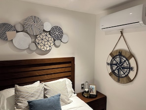 A bedroom with a wooden headboard, a bed with striped bedding, a nightstand with a framed photo and a decorative item, and a wall-mounted air conditioner.
