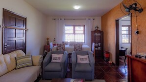 Egy nappali, amelyben van egy kanapé, két karosszék, egy étkezőasztal székekkel, egy szekrény és egy ventilátor.