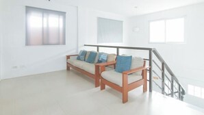 A spacious room with a wooden sofa set, a staircase, and large windows allowing natural light. - RedDoorz @ Qhouzz near PUP (Manila)