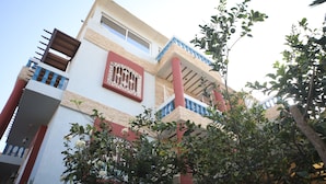 A multi-story building with balconies, a large window with decorative bars, and lush greenery in the foreground. - TAMRILUXE HOUSE (Tamri)