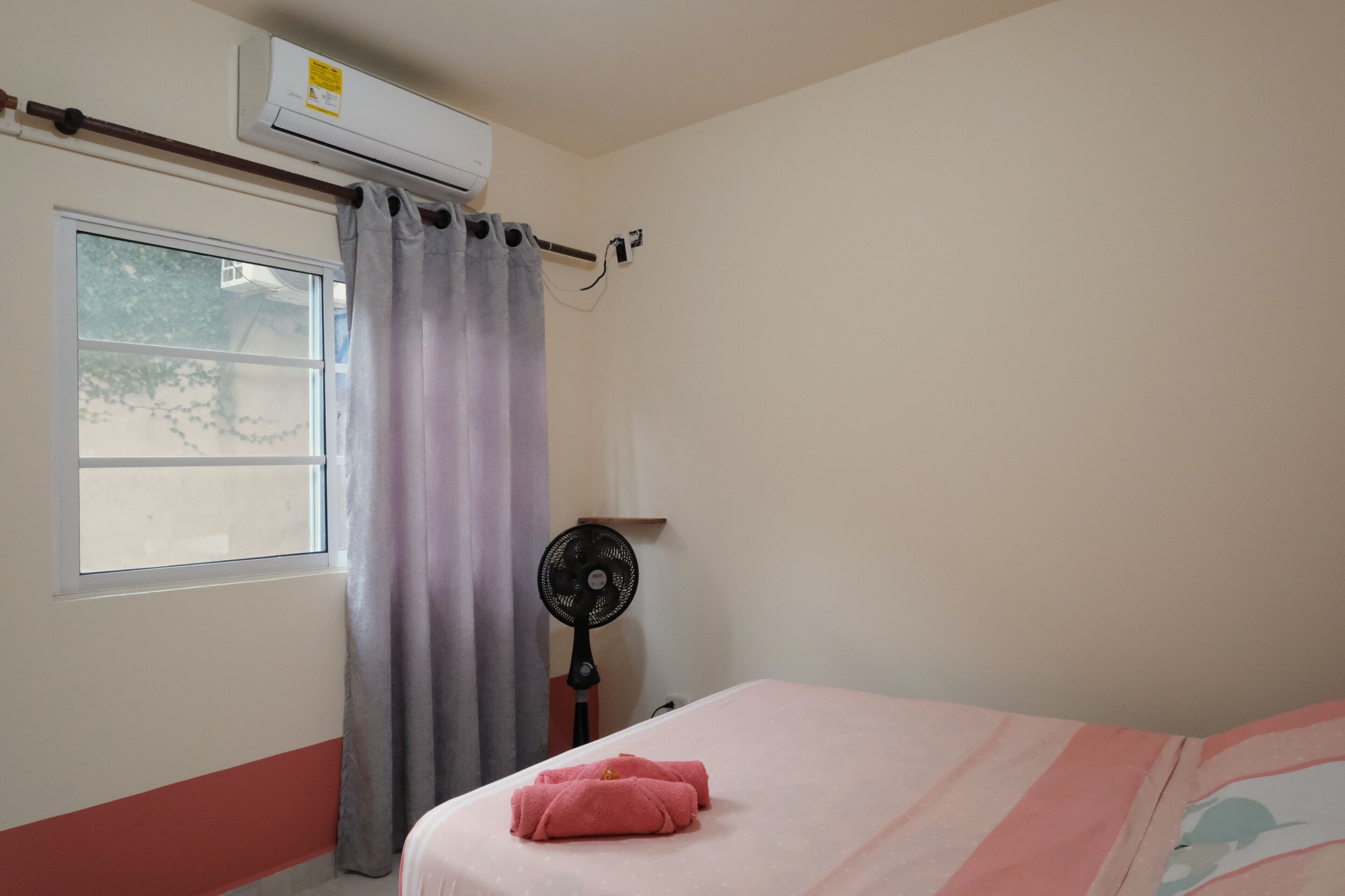 A room with a bed, a window with curtains, an air conditioner, and a standing fan.