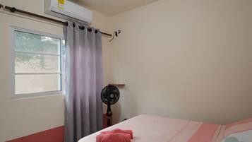 A room with a bed, a window with curtains, an air conditioner, and a standing fan.