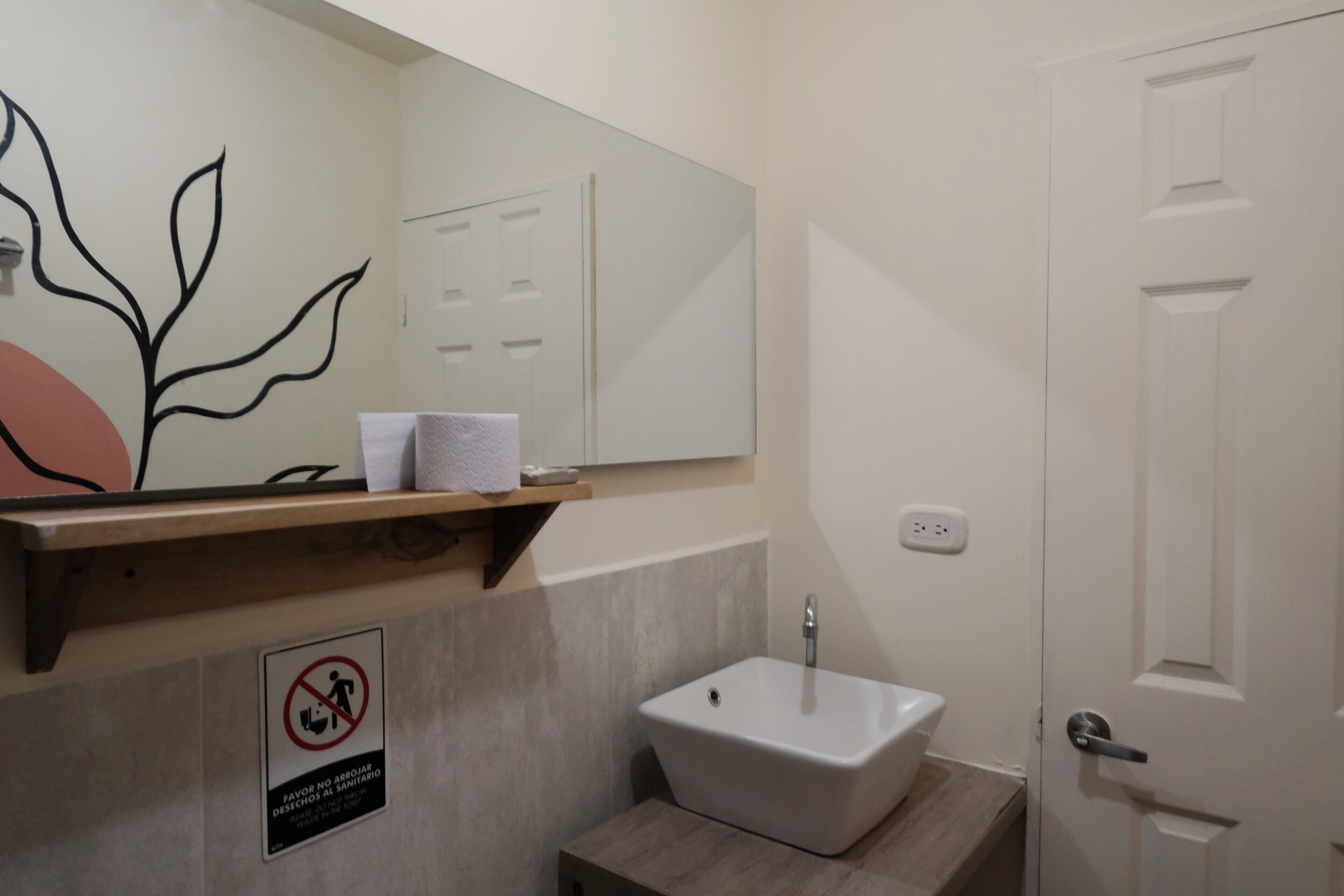 Un baño con lavamanos blanco, un espejo, dispensador de toallas y un cartel de prohibido fumar.