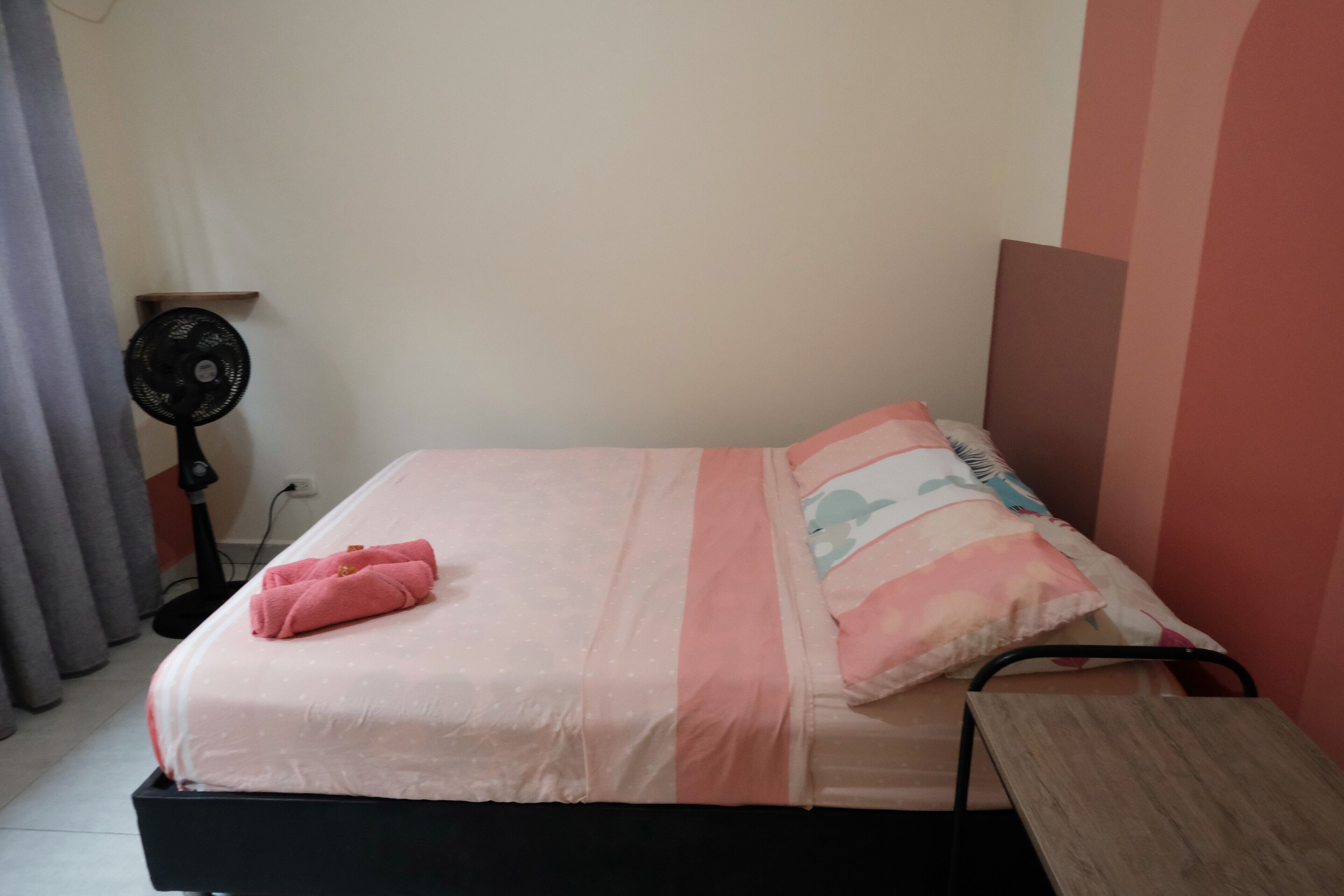 A single bed with a pink and white bedspread, a wooden bedside table, and a black fan.