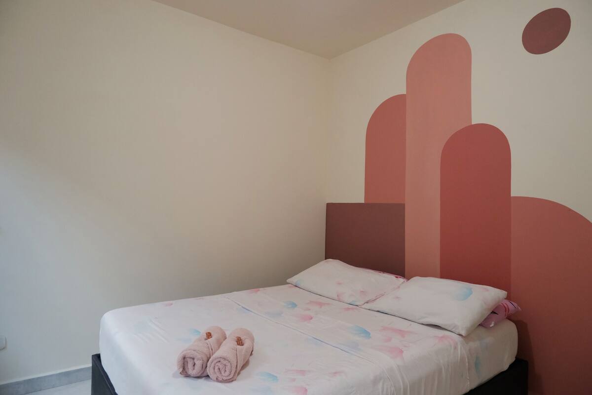 A single bed with a white and pastel-colored bedspread, a headboard, and two rolled towels on the bed.