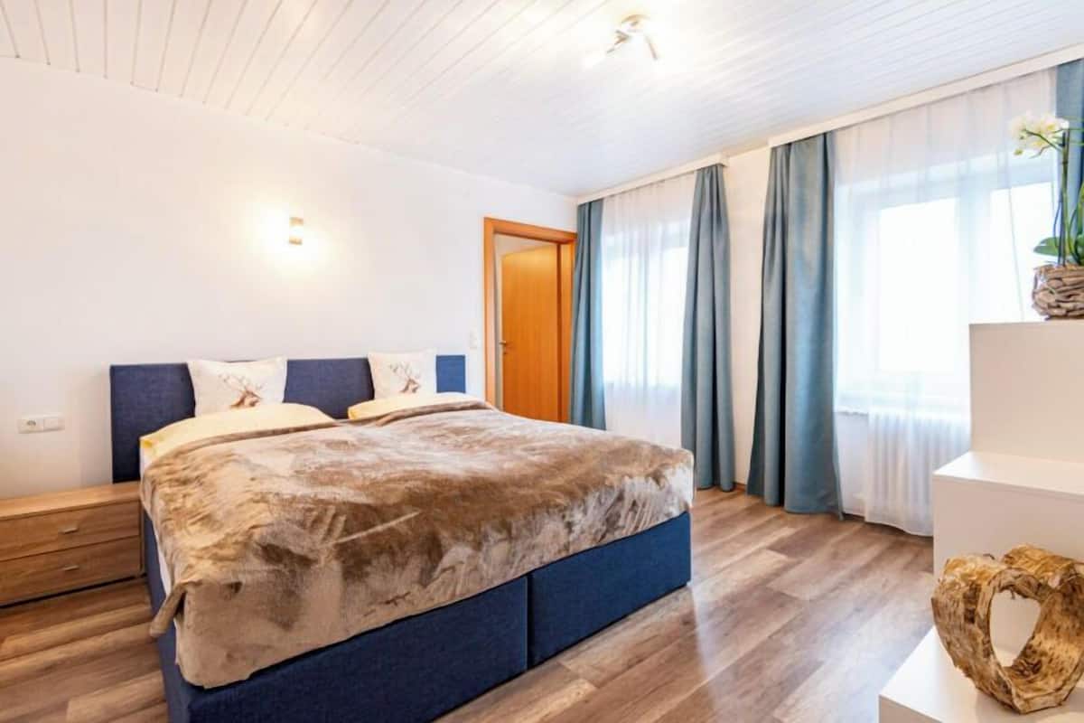 Une chambre avec un grand lit, un plancher en bois, une porte en bois et une fenêtre avec des rideaux.