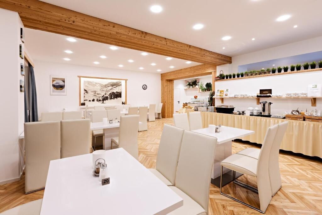 Un comedor moderno con mesas y sillas blancas, techo de madera y zona de cocina con estantes llenos de vajilla y plantas.