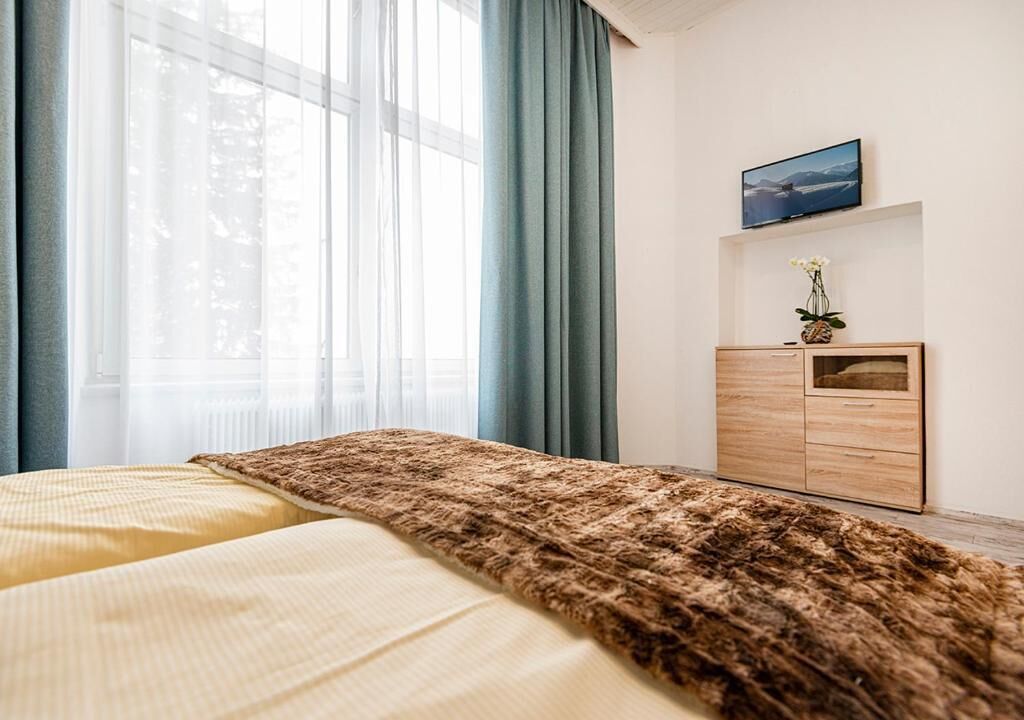A bedroom with a bed, a wooden dresser, a TV, and a window with curtains.