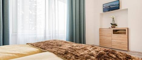 A bedroom with a bed, a wooden dresser, a TV, and a window with curtains.