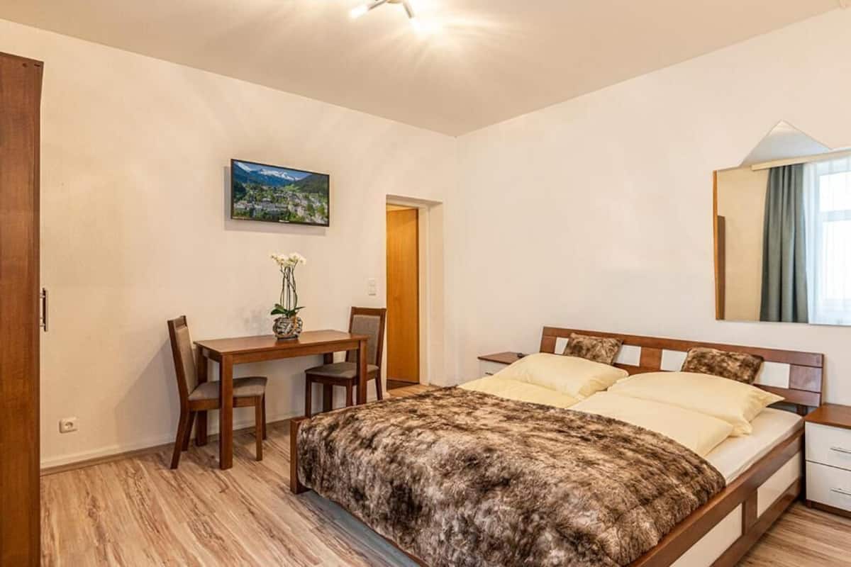 Une chambre à coucher avec un lit, une table de chevet et un bureau avec des chaises. Il y a un tableau au mur et une fenêtre avec des rideaux.