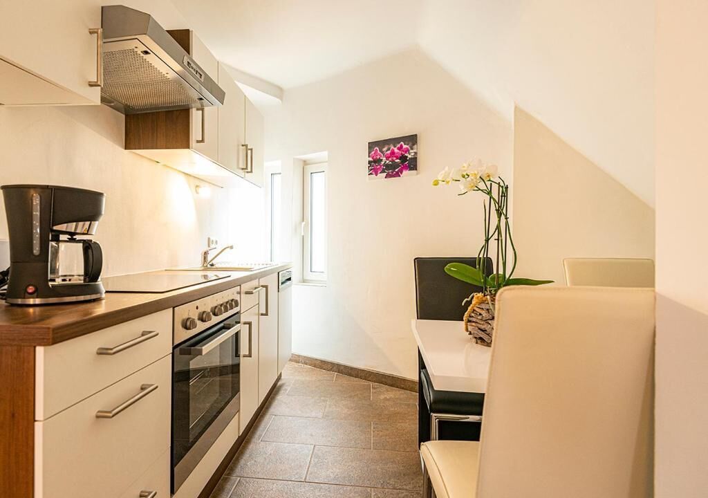 Cocina compacta con cafetera, horno y comedor con una silla y un florero.