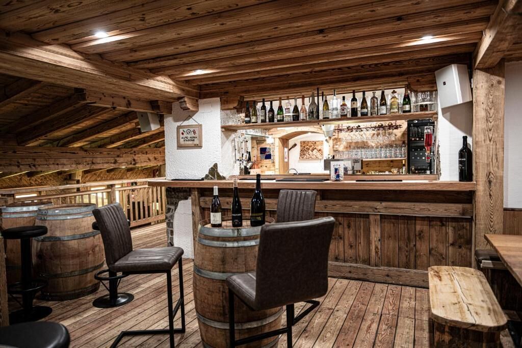 A rustic bar area with wooden furniture, bar stools, and a variety of bottles on shelves.