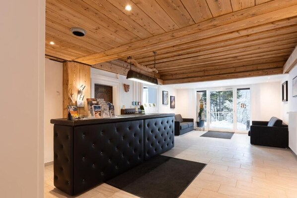 A modern lobby with a wooden ceiling, a black tufted reception desk, and a view of the outdoors through large glass doors.
