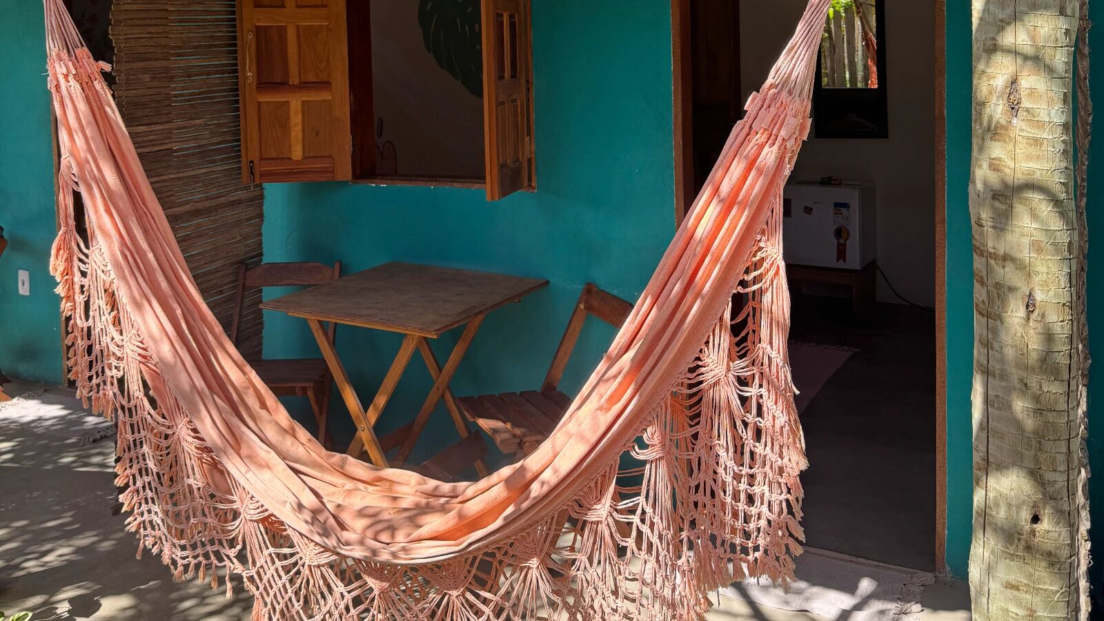 A hammock tied between two trees in front of a turquoise building with a wooden table and chairs inside.