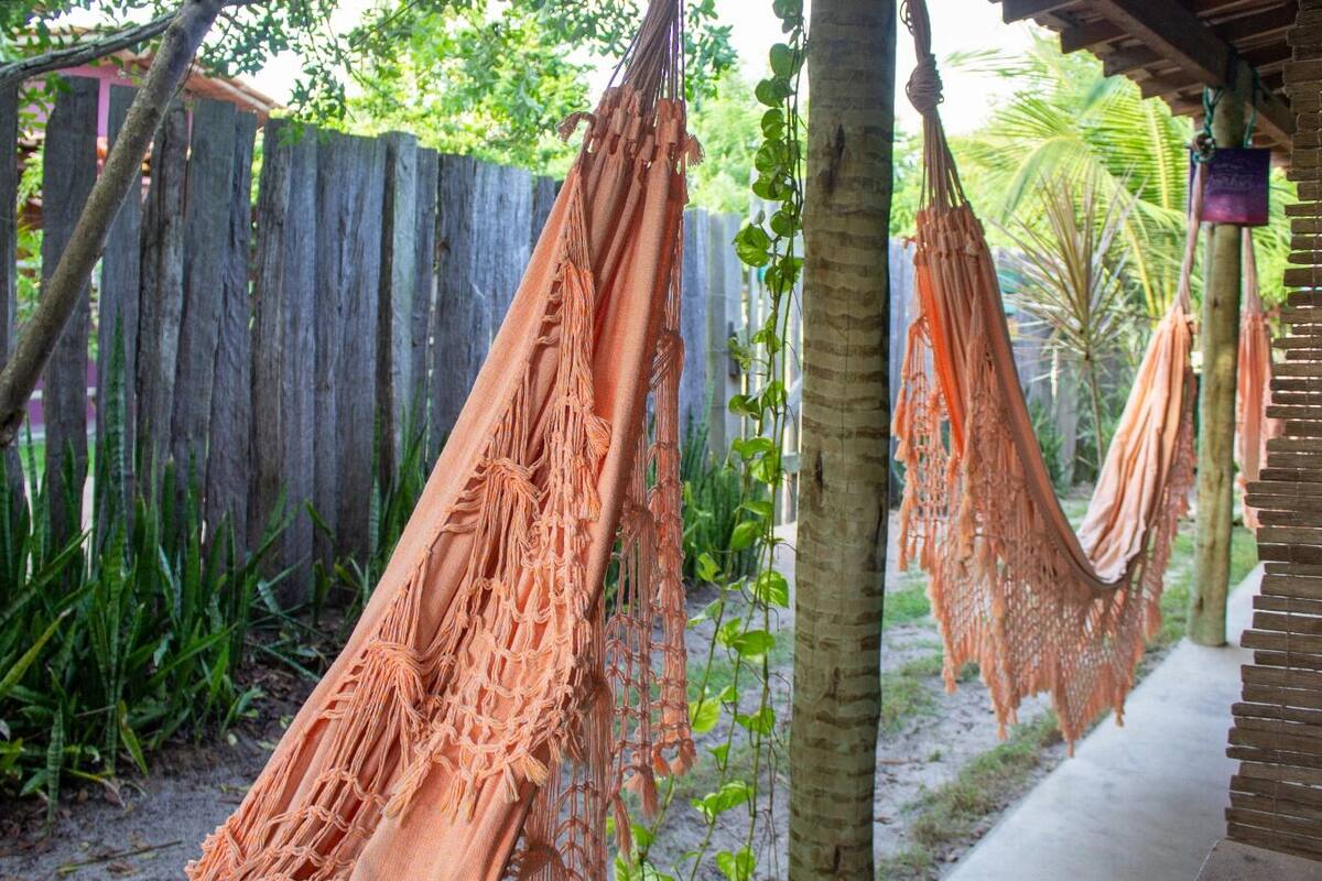 Un hamac est suspendu entre deux arbres dans un jardin, avec une clôture en bois et un chemin en béton.