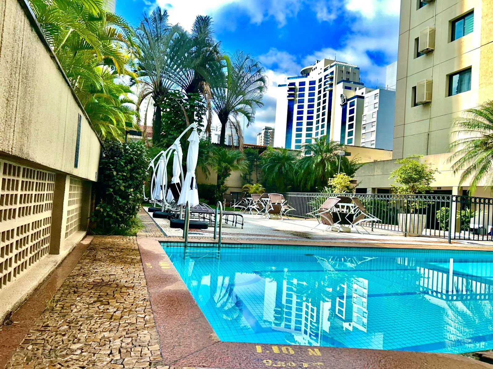 Uma piscina com espreguiçadeiras e guarda-sóis, cercada por palmeiras e edifícios modernos.