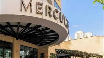 Um edifício com a palavra 'MERCUE' na fachada, uma área de entrada coberta e vista de prédios ao redor.