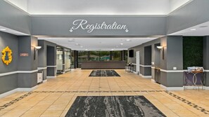 Een hotel receptie met een receptiebalie, een bord dat het registratiegebied aangeeft, en uitzicht op het buitengebied door grote ramen.