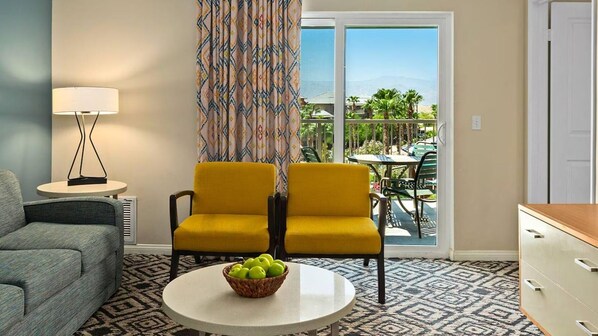 Ein Wohnzimmer mit einer Couch, zwei gelben Sesseln, einem kleinen Tisch mit Obstschale, einer Lampe und Blick auf einen Balkon mit Gartenmöbeln.