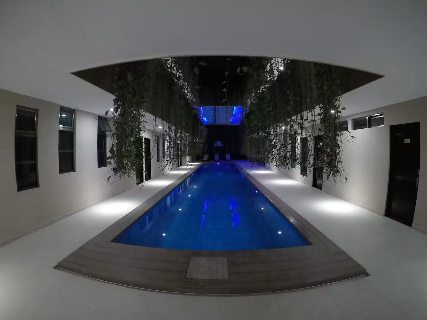 Piscina interior moderna com deck de madeira, rodeada por paredes brancas e grandes janelas.