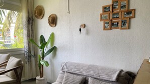 A cozy living room with a grey sofa, a wooden side table, a potted plant, and a wall-mounted light fixture. - T2 apartment with mezzanine in the Marina, 3 minutes from the beach (Les Trois-Îlets)