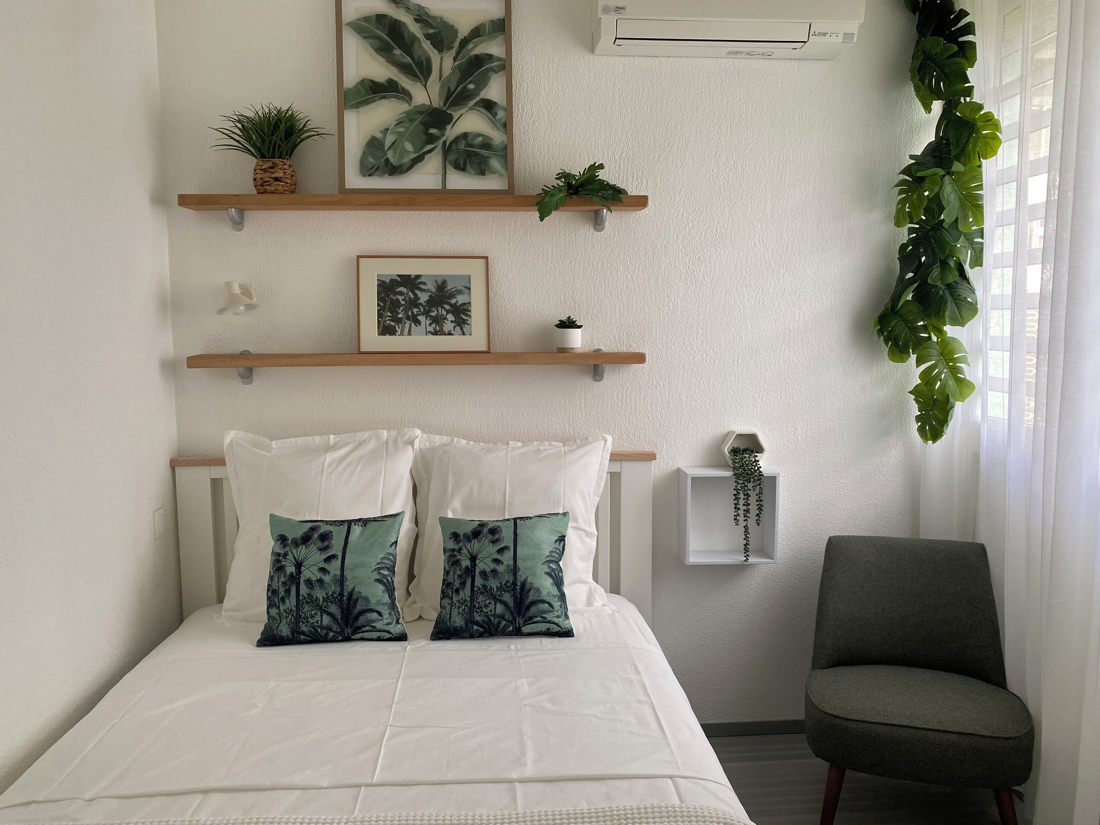 A bedroom with a bed, two pillows, a chair, a window with curtains, and wall-mounted shelves with plants and a picture.