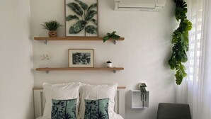 A bedroom with a bed, two pillows, a chair, a window with curtains, and wall-mounted shelves with plants and a picture. - T2 apartment with mezzanine in the Marina, 3 minutes from the beach (Les Trois-Îlets)