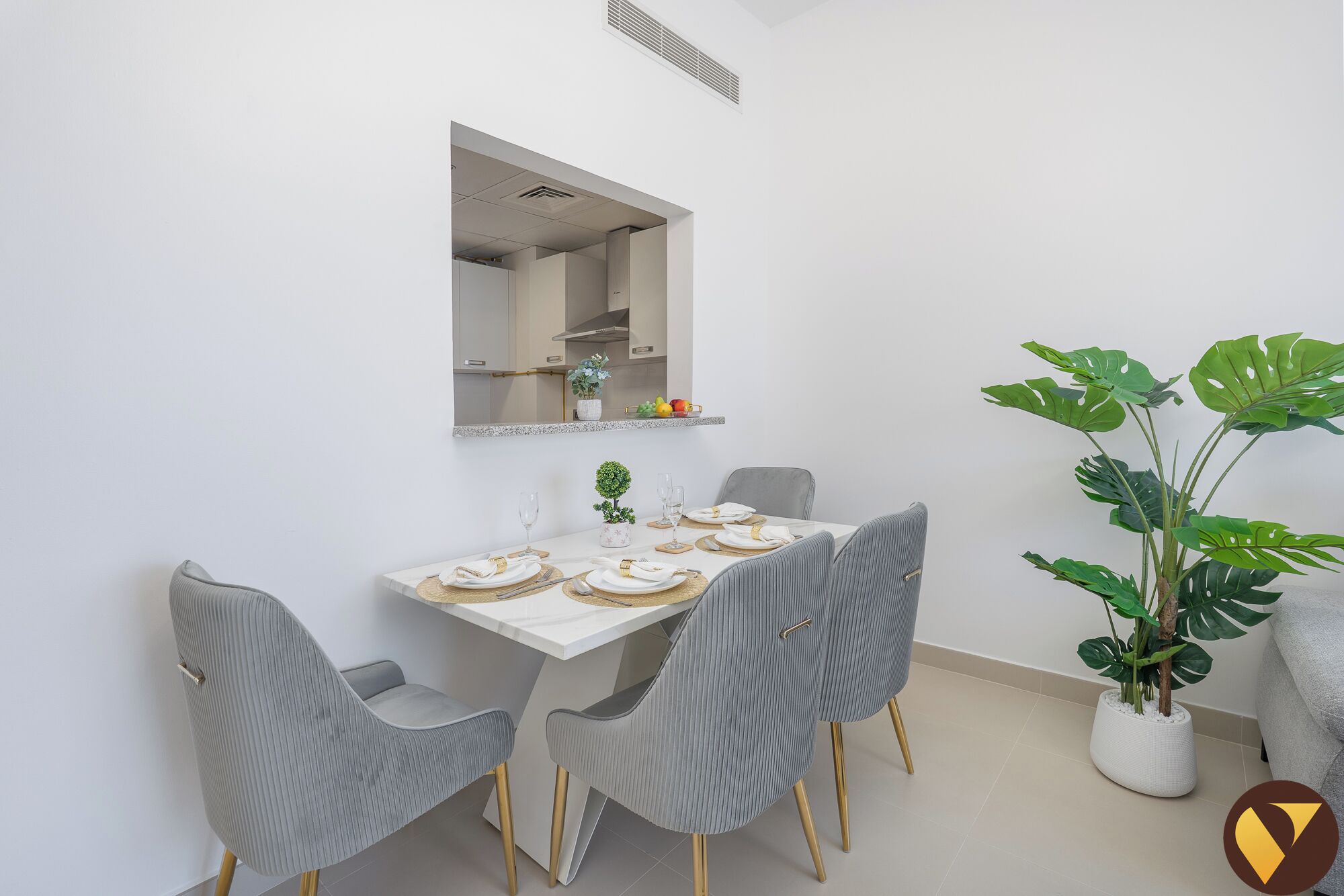 A dining area with a white table set for four, grey upholstered chairs with gold legs, a large potted plant, and a mirror above the table.
