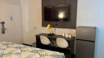 A hotel room with a bed, a desk with two chairs, a flat-screen TV mounted on the wall, a refrigerator, and a trash bin.