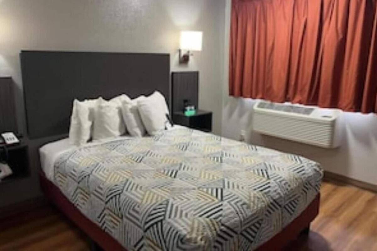 Ein Hotelzimmer mit einem großen Bett, einem Nachttisch, einer Lampe, einem Fenster mit Vorhängen und einer Wandklimaanlage.