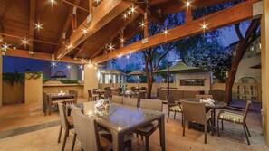Area patio esterna con travi in legno, luci a stringa e diversi posti a sedere, tra cui tavoli e sedie.