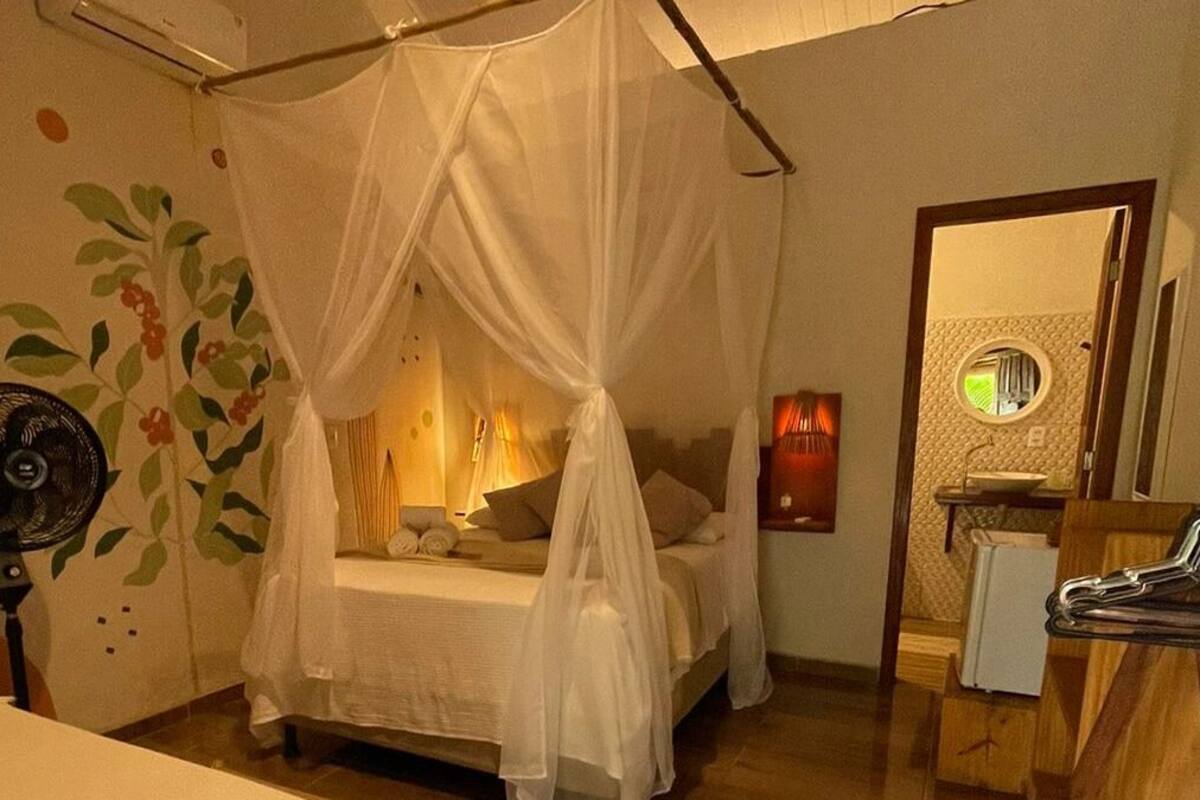 Een kamer met een bed onder een hemelbed, een nachtkastje met een lamp, een ventilator aan de muur en een spiegel.