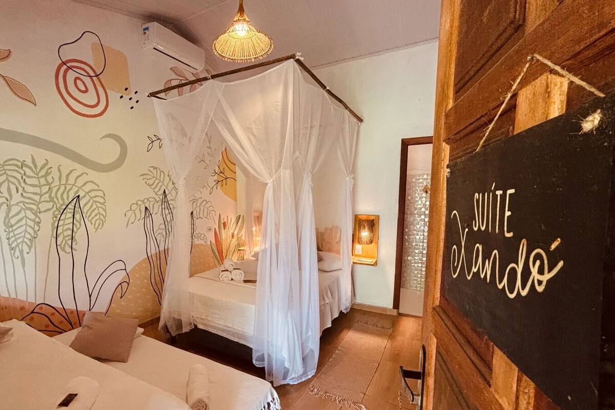 Een slaapkamer met hemelbed, muurschildering met plantenmotieven, houten wand met daarop 'SUITE Xando' en een hanglamp.