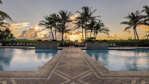 Un área de piscina con un paseo empedrado, sillas de descanso, palmeras y vista al atardecer.