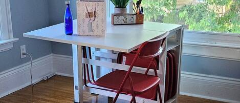 A room with a wooden floor, a white desk with a chair, a window with blinds, and a view of greenery outside.