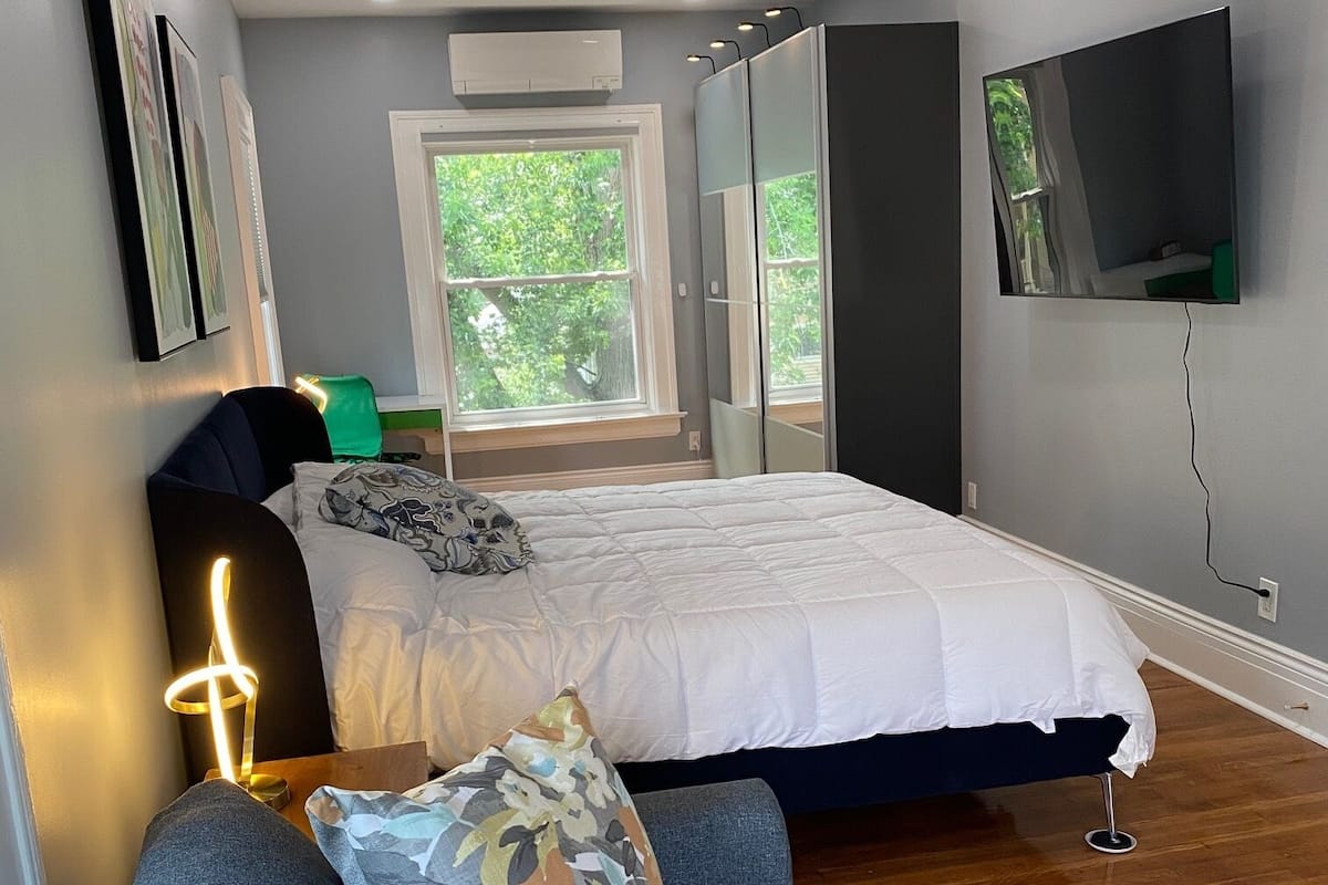 A modern bedroom with a bed, a ceiling fan, a TV, and a window with a view of trees.