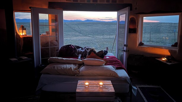 Ein gemütliches Zimmer mit einem Bett, einem Holztisch mit Kerzen und Blick auf ein Feld durch eine geöffnete Tür.