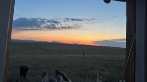 Ein Zimmer mit großem Fenster, das Blick auf ein grasbewachsenes Feld und Berge im Abendlicht bietet.