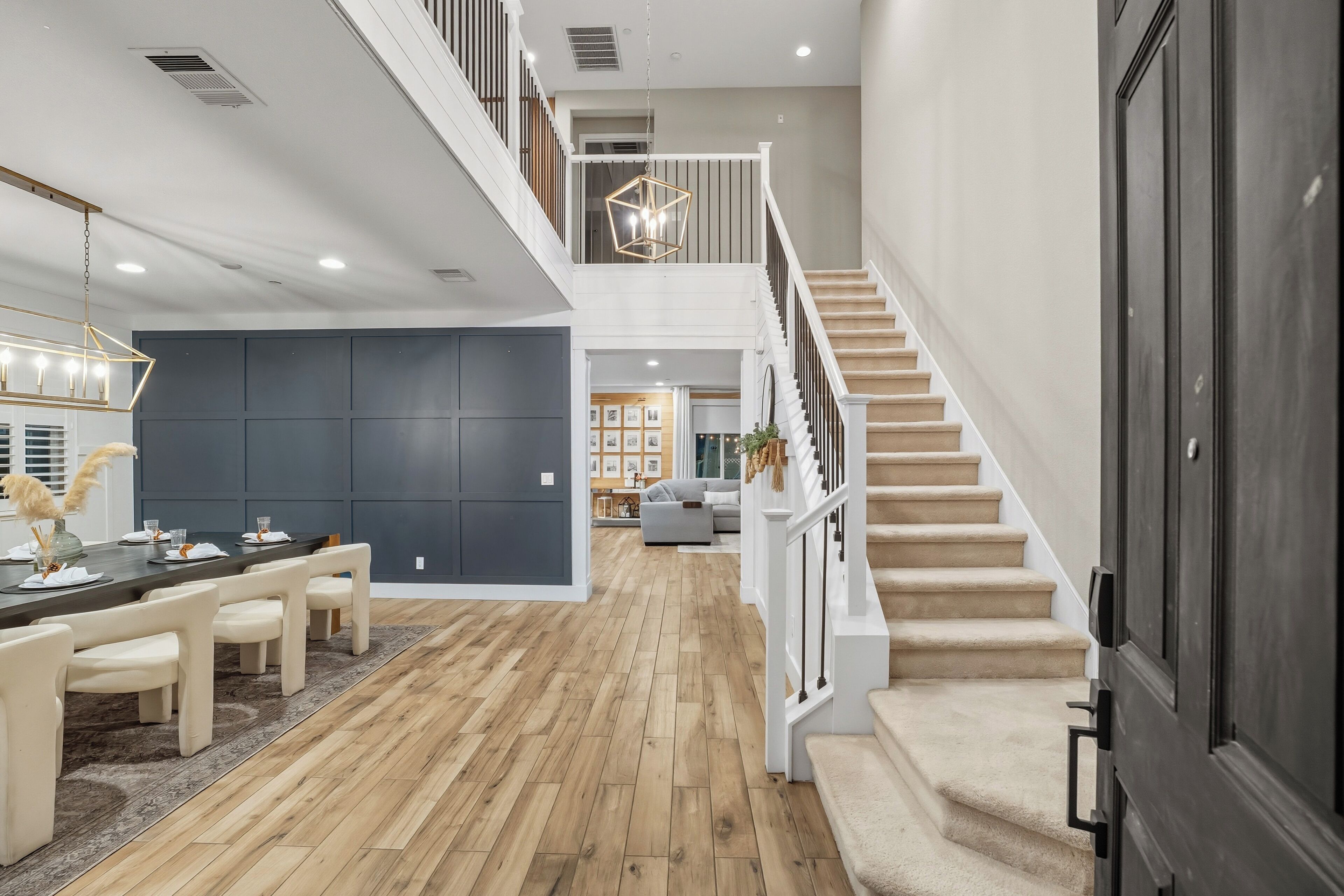 A spacious living area with a wooden floor, a dining table with chairs, a staircase, and a modern chandelier.