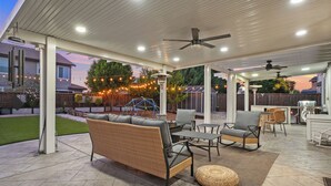 An outdoor patio with a covered seating area, including a sofa and chairs, a dining table, and a ceiling fan.