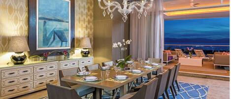 A dining area with a long table set for a meal, a chandelier, and a city view through large windows.