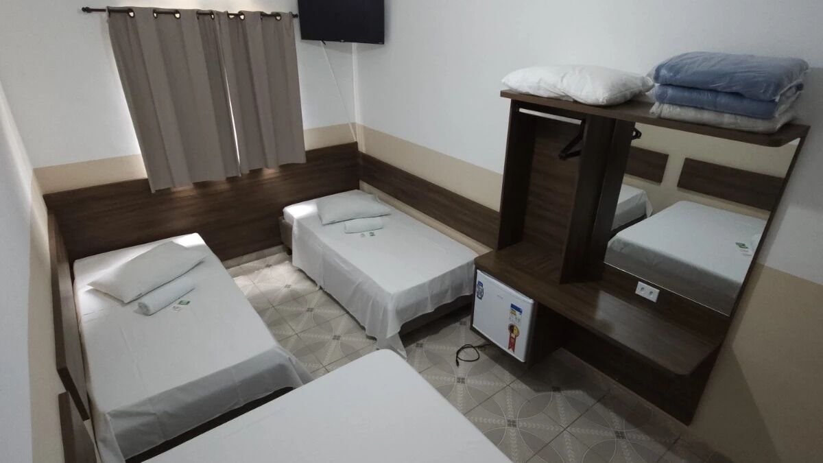 Quarto com três camas de solteiro, uma televisão na parede e uma pequena mesa com toalhas dobradas.