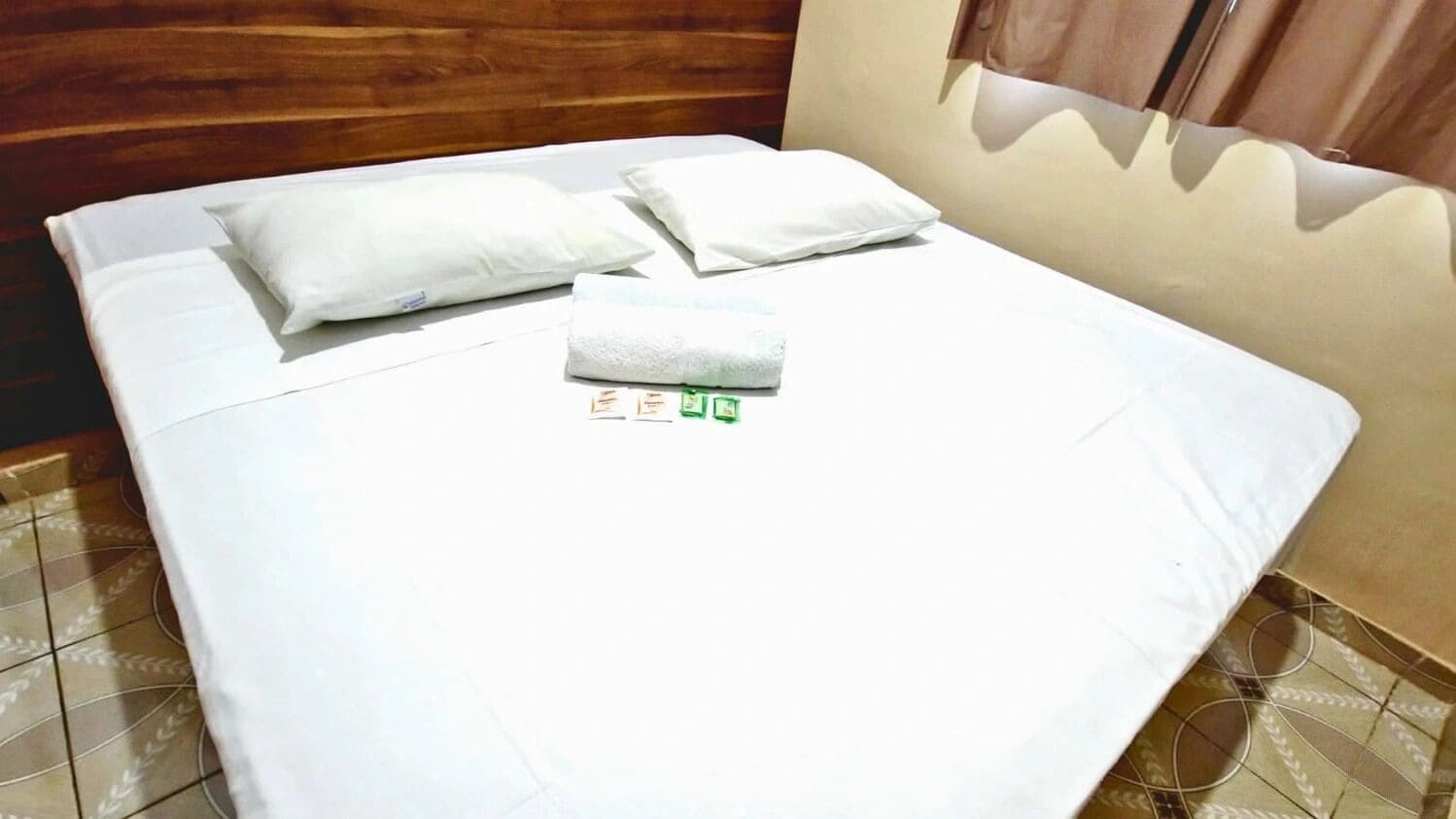 A neatly made bed with white linens and pillows, a folded towel, and a small green and white item on the bed.