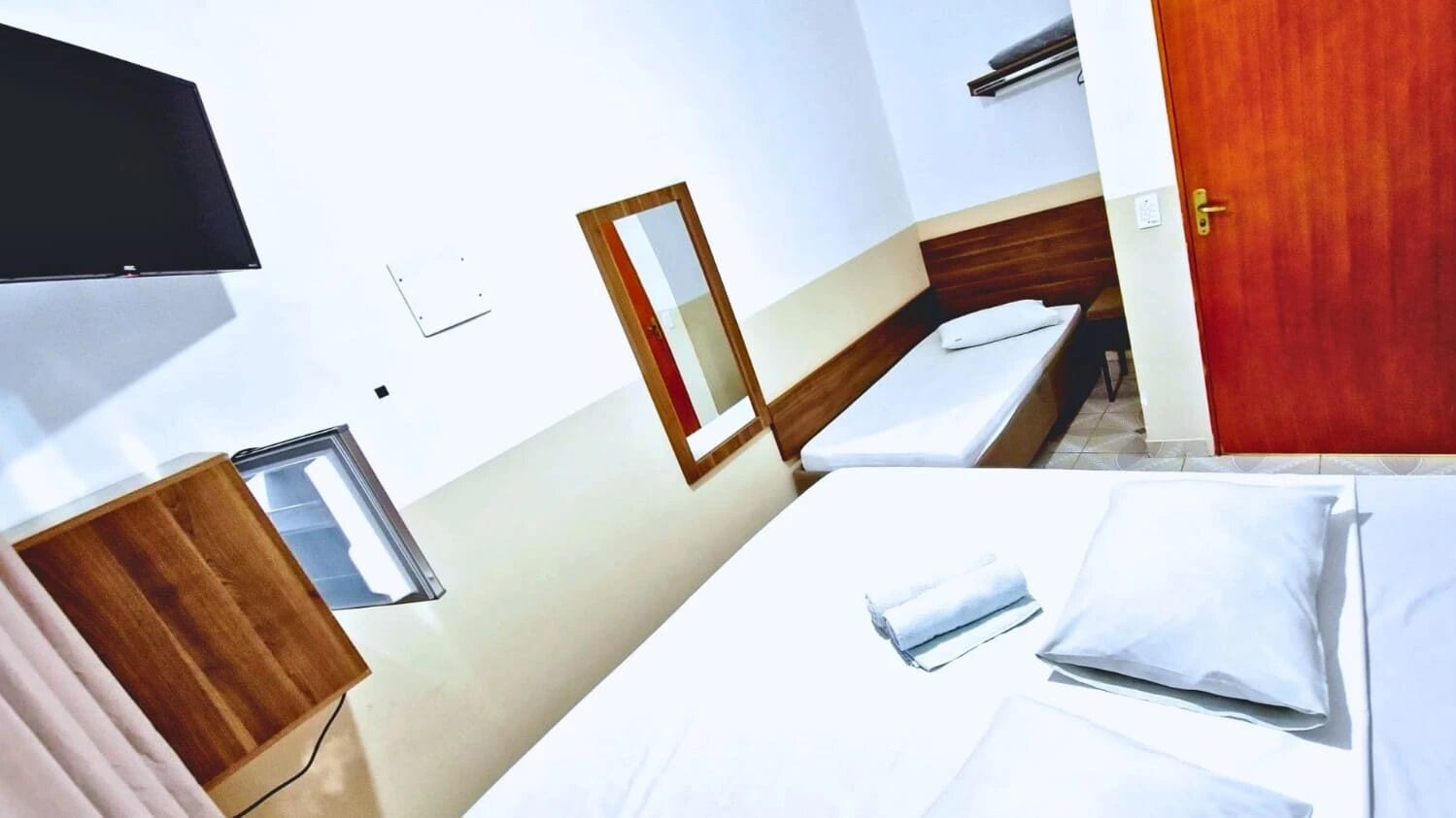 Una habitación de hotel con cama, cabecera de madera, mesita de noche, espejo y televisor fijado a la pared.